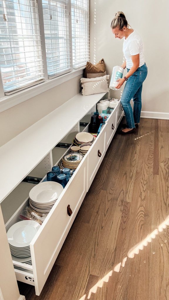 The picture shows storage bench seating. A woman is putting away kitchen appliances and you can see that they have put in plates, glasses and cleaning supplies